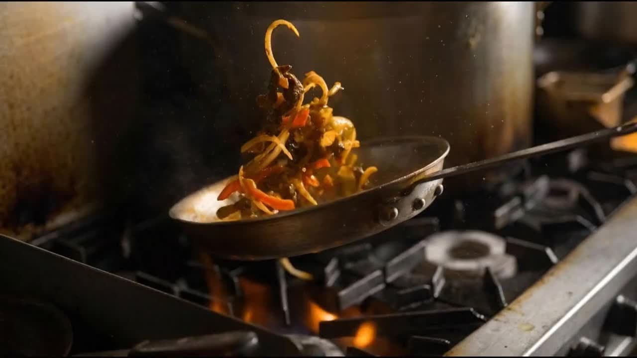 Sliced peppers, onions and meat tossed in a skillet above a gas flame on a commercial kitchen stove